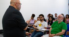 Na fotografia, um professor branco se encontra à esquerda. Ele está de frente para diversos estudantes sentados em carteiras.