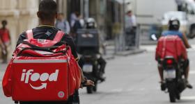 Foto de entregadores de aplicativo em uma rua. No lado esquerdo, uma pessoa negra, de cabelo raspado e de costas, pedala uma bicicleta. Ela usa camiseta preta e uma grande mochila vermelha em formato quadrado, a bag, com o logo do Ifood. Em segundo plano, um pouco desfocados, duas pessoas em motos: a do lado direito tem a pele parda, usa capacete e bermuda pretos, regata azul e bag  vermelha. A pessoa à  esquerda usa capacete e calça pretos e tem o resto do corpo coberto pela bag, que é preta.