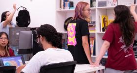 Foto de estudantes, todos brancos em uma sala. À esquerda, duas pessoas estão sentadas em uma mesa e operam computador e laptop, enquanto, à direita, duas pessoas conversam em pé. Atrás delas, há uma restante repleta de livros e outros objetos. Ao fundo, do lado esquerdo, uma esudante mexe em uma mochila preta sobre um armário.