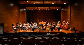 Foto de uma orquestra de câmara em um palco. São cerca de 30 jovens músicos sentados e distribuídos em semicírculo, a maioria com instrumentos de cordas. O regente está sentado em uma cadeira sobre um tablado e tem os braços erguidos, com a batuta na mão direita. Ele está de costas para plateia, que está praticamente vazia. Todos usam roupas coloridas e informais.