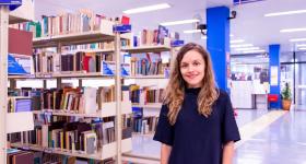 Fotografia de Lilian Viana, mulher branca de cabelos longos e loiros, que veste uma blusa azul escuro e usa um relógio no pulso direito. Ao fundo, há uma biblioteca com prateleiras repletas de livros. Há uma placa no canto superior direito escrito: Livros.