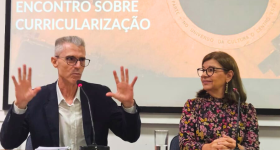 Foto em plano médio. À esquerda, homem branco, de cabelos curtos e grisalhos, usando óculos de grau de haste escura e uma camisa branca quadriculada e um blazer preto. Ele está sentado, ergue as duas mãos para cima, na altura do ombro, enquanto fala ao microfone. Ele é observado por uma mulher que está sentada à direita. Ela é uma mulher branca, de cabelos castanhos, lisos e na altura dos ombros. Ela usa óculos de haste escura e arredondada, brincos dourados, e um vestido preto de manga longa com estampa.