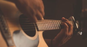 Foto das mãos de uma pessoa tocando um violão de cores claras. Uma das mãos segura o braço do violão, enquanto a outra dedilha as cordas. O fundo é escuro.