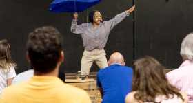 Foto de uma performance em que uma pessoa está com uma máscara dourada de rosto triste. Com a mão direita ela segura um guarda-chuva azul e estende a esquerda na diagonal alta. Ela usa camisa cinza, calça bege e se apresenta diante de uma pequena platéia de costas para a foto. O cenário é preto.