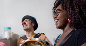 Na foto, um homem branco à esquerda e uma mulher negra à direita tocam instrumentos e cantam sorridentes. O homem usa chapéu azul, nariz de palhaço vermelho e blusa laranja listrada. A mulher, que está em primeiro plano, tem cabelos cacheados castanhos até a altura dos ombros, usa óculos de armação preta e camiseta preta. Ela segura um pandeiro que encobre o instrumento dele e ambos estão voltados para o lado esquerdo da imagem. Ao fundo, uma parede branca e um armário, sobre o qual há alguns potes plástico