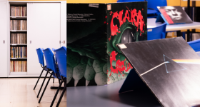 Foto de capas de discos em vinil dispostas sobre uma mesa. No canto inferior direito, parte de uma capa mostra, sobre um fundo preto, a imagem de um feixe colorido que sai de um prisma triangular. Ao centro, uma capa de fundo preto com destaque para um olho cercado de detalhes verdes e escamas e dos dizeres, em vermelho, Clara Crocodilo. Ao fundo, cadeiras plásticas azuis, um armário branco e uma parede amarela.