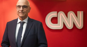 Foto de um homem branco sorrindo. Ele veste terno e gravata azuis, óculos de armação quadrada e preta. Ao fundo, uma parede vermelha e um letreiro com o texto “CNN”.