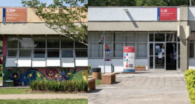 Montagem com duas fotografias de fachada de prédios. À esquerda, o Departamento de Relações Públicas, Propaganda e Turismo. No alto, há uma placa laranja escrito CRP e, abaixo, janelas de vidro e uma parede com grafite. À frente um caminho de bloquetes, grama e plantas.  À direita, a fachada do Departamento de Jornalismo e Editoração. No alto, há uma placa vermelha escrito CJE e, abaixo, uma porta e janelas de vidro. À frente do prédio há um totem de informações, uma cesta de lixo e bancos. 