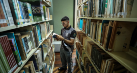 Foto de um rapaz folheando um livro em uma biblioteca.  Ele usa óculos, tem cabelos curtos e pretos, veste roupa esportiva e está entre dois corredores de estantes cheias de livros.