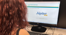Fotografia de uma mulher olhando a tela de um computador. A mulher está de costas para a câmera, tem pele branca e cabelo vermelho e observa a tela do computador onde é há uma logo escrita “Júpiter web”. 