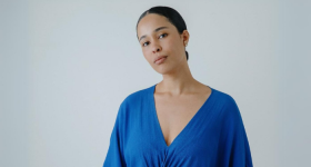 Na foto, uma mulher negra com cabelos pretos presos em um coque baixo. Ela veste blusa azul com decote V e usa brincos. O fundo é cinza.