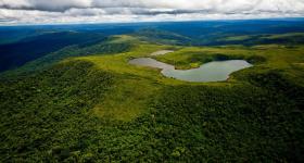 Foto panorâmica da Floresta Nacional de Carajás vista de cima. Em primeiro plano, há um grande morro coberto por vegetação e, no topo dele, um lago. Em segundo plano, há um mar de morros também cobertos por vegetação, sendo que quanto mais distante estão, mais escuros e azulados. Acima da linha do horizonte, o céu nublado. 