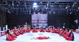 fotografia de um grupo de pessoas sentadas em um palco na forma de um semicirculo, com uma pessoa deitada de barriga para baixo no centro da roda. Todos usam saias compridas e blusas vermelhas e têm etnias variadas. O palco possui algumas cadeiras brancas e instrumentos musicais ao fundo e, no teto, há muitos holofotes. 
