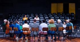 Fotografia de um grupo de doze pessoas sentadas em cadeiras de madeira em semicírculo no palco de um teatro. De frente para elas está a plateia com apenas duas pessoas sentadas. As pessoas nas cadeiras estão de costas para a câmera e têm diversas aparências e cores de vestimenta. O piso é de madeira, as paredes do teatro são escuras e, ao fundo, há duas portas de incêndio amarelas fechadas.