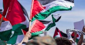 Foto de uma manifestação na Palestina. No centro, é possível ver o braço de uma pessoa segurando um mastro com uma bandeira da Palestina, que tem uma linha horizontal preta, uma branca e uma verde e um triângulo vermelho à esquerda. À esquerda e à direita há mais pessoas segurando a mesma bandeira. Ao fundo, há um céu azul.
