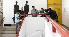 Foto de um grupo de sete pessoas que conversa apoiado em um corrimão vermelho em formato de U. No centro, se destaca o vão criado pela escada, que aparece à esquerda. À direita, um corredor e uma porta amarela. Ao fundo, uma parede branca com reproduções em grande formato de fotos em preto e branco de duas pessoas. 
