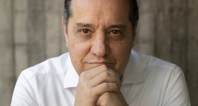 Foto de um homem branco, calvo, de cabelos escuros e curtos. Ele veste camisa social branca e relógio de pulso prateado. Sorri ligeiramente, com as mãos unidas sob o queixo e os cotovelos apoiados sobre uma mesa. O fundo é cinza e desfocado.