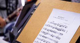 Foto da partitura da música Rondido. A página impressa está apoiada num suporte de madeira com um metrônomo apoiado no canto inferior esquerdo. Ao fundo, desfocada, do lado esquerdo, há uma pessoa tocando um instrumento de sopro.