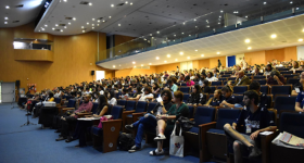 Foto de diversas pessoas sentadas na plateia de um grande auditório. O chão tem carpete azul e as paredes são de madeira clara. No teto branco há diversas lâmpadas amarelas e brancas. Alguns microfones estão posicionados em pedestais diante da primeira fileira de assentos.