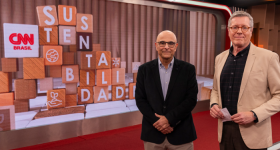 Foto de dois homens brancos à frente de uma tela digital na qual aparece o logo CNN Brasil e letras pintadas em blocos de madeira empilhados formando a palavra Sustentabilidade. O homem à esquerda é careca, usa óculos de armação quadrada e preta, camisa azul, blazer preto e calça marrom. O homem à direita usa camisa preta, blazer bege, calça preta, óculos de armação redonda e marrom e tem cabelos curtos e grisalhos. 