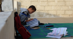 Fotografia de um menino em perfil escrevendo em um caderno. Ele é branco, de cabelos castanhos e curtos, veste um agasalho cinza, tem o olhar voltado para o caderno e está sentado em um tapete verde e azul, no chão, com pernas cruzadas, com o caderno no colo. À frente dele, há uma mochila vermelha e um livro aberto, no chão. Atrás dele, há uma parede cinza com tijolos aparentes. 