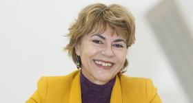Foto de uma mulher branca, de cabelos loiros, curtos e lisos. Ela veste uma blusa roxa e um paletó amarelo. Ela está com as mãos uma sobre a outra e apoiadas sobre uma mesa. O fundo é branco.