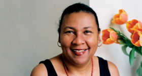 Foto de mulher negra de olhos castanhos e cabelos escuros e presos sorrindo. Ela veste regata preta, usa colar de contas vermelhas e brincos de argolas douradas. No fundo, uma parede cinza e branca e flores cor de laranja à direita.