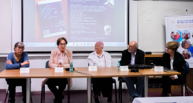 Foto de um grupo de cinco pessoas. Todos são brancos e apenas um é homem. Eles estão sentados atrás de uma mesa de madeira, na qual há plaquetas com os nomes de cada um, microfones e garrafas d'água. No fundo, uma projeção na parede com textos e imagens. Também há um banner no canto direito com fotos e o título “Comunicação & Educação” em azul. 