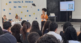 Foto de uma mulher negra falando ao microfone para um grupo de pessoas sentado à sua frente e de costas para a câmara. Ela está de pé, tem cabelos crespos que estão presos, usa uma camiseta laranja e uma calça preta. Atrás dela, uma mesa com duas mulheres e um painel branco onde está escrito “De volta à Escola”, “Eu na USP”, decorado com várias estrelas, em amarelo e azul. Atrás dela também há uma televisão que mostra um código QR. 
