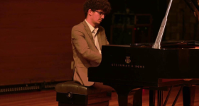 Foto de um homem negro de pele clara, sentado em um banco e tocando piano de cauda. Ele tem cabelos crespos, castanhos e curtos, veste um terno marrom com camisa branca e usa óculos de armação preto. O fundo é escuro.