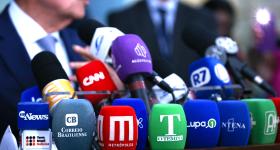 Foto de um homem concedendo uma entrevista com vários microfones a sua frente. Ao fundo há dois homens: à esquerda um homem branco com terno preto, camisa branca e gravata estampada em roxo e azul; à direita, um homem, com terno e gravata pretos e camisa branca. Seus rostos estão cortados. Na frente deles há uma mesa com diversos microfones coloridos com logomarcas de veículos de comunicação.