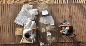 Foto de cima de duas mulheres sentadas em cadeiras sobre um piso de madeira. Ambas são brancas, de cabelos castanhos, mas uma delas aparenta ser mais jovem que a outra. Entre elas, há uma rede de pesca com papéis escritos dispostos sobre ela. Ao lado da mulher sentada à esquerda, um grande tecido claro coberto por escritos em azul.