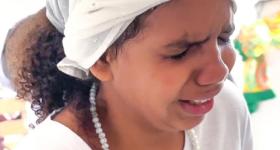 Foto de menina negra de olhos fechados e com expressão emocionada. Ela veste um turbante branco, tem cabelos castanhos escuros cacheados na altura do ombro e usa um fio de contas brancas e camiseta branca. O fundo é claro. 
