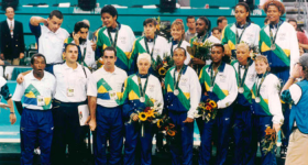  Foto da seleção brasileira feminina de basquete posando com medalhas de prata. Do lado esquerdo, o técnico e outros homens da comissão técnica. Os homens vestem calças pretas e azuis e camiseta branca com detalhes em verde, amarelo e azul. As mulheres vestem calça azul e agasalhos brancos com detalhes em verde, amarelo e azul. Uma pessoa à frente segura uma câmera de vídeo e veste bermuda bege e camiseta azul. Ao fundo está uma arquibancada e algumas pessoas.