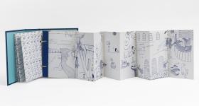 Foto de livro com capa azul e páginas brancas que se abrem em sanfona. Nas páginas, há diversos desenhos aparentemente feitos em caneta azul. O fundo é branco. 