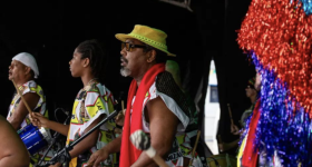 Foto de um grupo de pessoas negras de perfil, sendo que três estão em foco e seguram baquetas e tambores. Elas usam roupas coloridas de branco, amarelo, preto, vermelho e verde. Uma pessoa usa boina branca e outra um chapéu amarelo. O fundo é escuro e, no canto direito da imagem, há franjas vermelhas e azuis de papel celofane. 