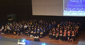 Foto de cerca de 150 estudantes, vestindo becas e sentados lado a lado em cadeiras posicionadas no palco de um auditório. Ao fundo, uma tela de projeção, que exibe arte em que se lê, parcialmente, “Colação de grau da ECA-USP”. À frente do palco, há três arranjos de flores.