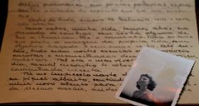  Em uma superfície, uma carta com escritos em letra cursiva e tinta preta e uma fotografia em preto e branco de uma mulher sobre a carta, no canto direito da imagem. A mulher tem cabelos curtos ondulados e sorri olhando na direção de algo que não aparece na foto. A carta é amarelada e a fotografia está com manchas amareladas também. 