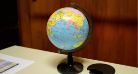 Foto de um pequeno globo terrestre que mostra principalmente o continente asiático, com países nas cores amarelo, rosa, laranja e verde, e o oceano Índico em azul. O globo está sobre uma mesa clara com detalhes em preto. Sobre a mesa há também um papel colorido. O fundo, desfocado, é uma porta de madeira. 