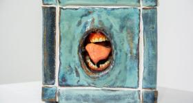 Foto de escultura em cerâmica de um cubo com uma boca aberta no centro. O cubo é azul e sua estrutura lembra pisos de cerâmica com rejunte branco. A boca é um buraco no centro do cubo e exibe língua, dentes e gengivas. 