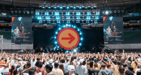 Foto de show musical com centenas de pessoas. A maioria do público é jovem e veste roupas claras. No palco, a banda e, ao fundo, um cenário com refletores e a imagem de uma seta vermelha em um círculo laranja, apontando para a direita. Acima, refletores com a cor azul. O palco é ladeado por telões que exibem imagens da banda.