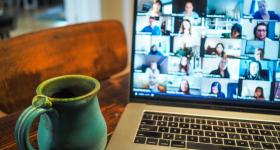 Foto de notebook sobre uma mesa. Na tela, há diversas imagens de pessoas em uma reunião online, ao lado do computador há uma caneca verde. 