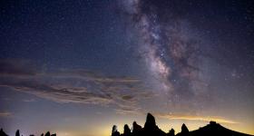 Foto de céu estrelado no fim da tarde. Na parte superior, há um aglomerado de estrelas brancas, com o céu azul escuro, na parte inferior, há um terreno pedregoso e quase totalmente escuro. O fundo é azul escuro.