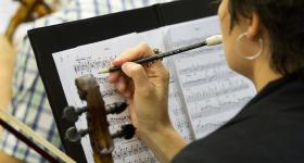 Mulher branca, de cabelos curtos e usando um brinco de argola, segura um lápis enquanto faz anotações em uma partitura. Segura também um violino. Ao fundo, outros músicos. 