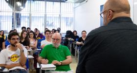 Foto de uma sala de aula. Um homem branco, calvo, usando oculos e blusa preta, está de costas. Um grupo de pessoas, sentadas em carteiras escolares, está a sua frente, e o observa. Ao fundo, janelas e uma parede branca.
