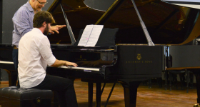 Foto de uma aula de piano. Um jovem, de cabelos castanhos e barba, está sentado de costas, com um piano preto a sua frente e tocando o instrumento. Ele veste uma camisa branca, calça preta e tenis. Ao seu lado, está um homem branco, de cabelos curtos e grisalhos, usando óculos e uma camisa azul e branca listrada. Ele observa as suas duas mãos, enquanto conversa com o jovem. O chão é de madeira e ao fundo uma parede preta e a vista parcial de um projetor