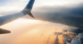 Foto da asa direita de um avião. A asa está posicionada no canto esquerdo da foto. No horizonte há nuvens contornadas por diferentes tons de laranja e sombreados de um sol poente.