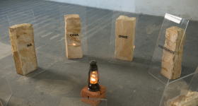 Foto de lampião aceso sobre um pedestal com diversas pedras em semicírculo ao seu redor. As pedras são claras e na frente de cada uma há uma placa transparente com os dizeres, da esquerda para a direita: “na”, “casa”, “onde”, “moramos”. O fundo é cinza claro. 