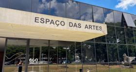 Foto de um edifício com janelas de vidro escuro espelhado e um letreiro escrito Espaço das Artes em uma estrutura de concreto que serve de cobertura sobre a entrada. Acima do edifício, o céu está azul com algumas nuvens brancas. O reflexo dos vidros mostra grama e árvores verdes, carros, pessoas e outros prédios. 
