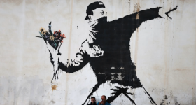 Foto de grafite na cor preta sobre um muro claro. O desenho retrata um homem com boné e rosto coberto segurando um buquê de flores, em um gesto de quem se prepara para lançar o objeto, semelhante ao gesto de quem lança uma granada ou um coquetel Molotov. As flores são amarelas e vermelhas.